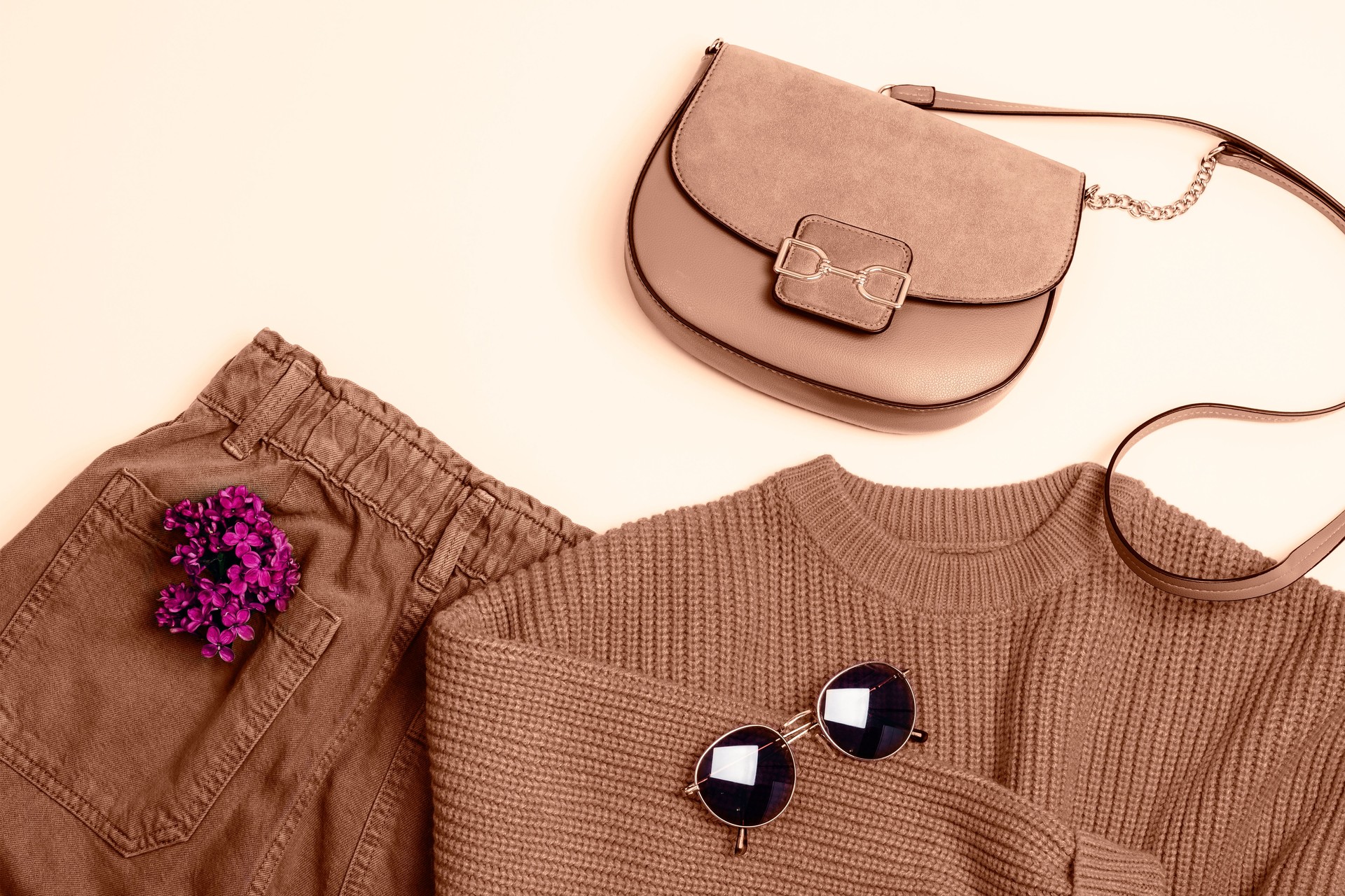 Stylish woman's outfit: knitted sweater, jeans, sunglasses and handbag. Lilac flowers decoration. Top view. Demonstrating color of the year 2025 - Mocha Mousse
